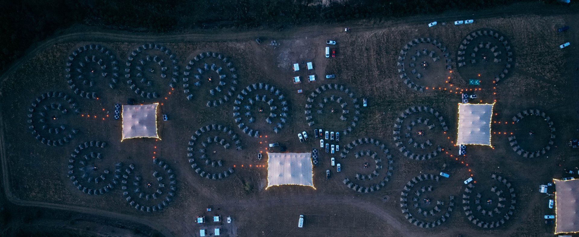A Burn Camp at dusk, seen from directly above: spirals of tents around their fires, linked by lantern-lit paths to the glowing Lapa Lounges