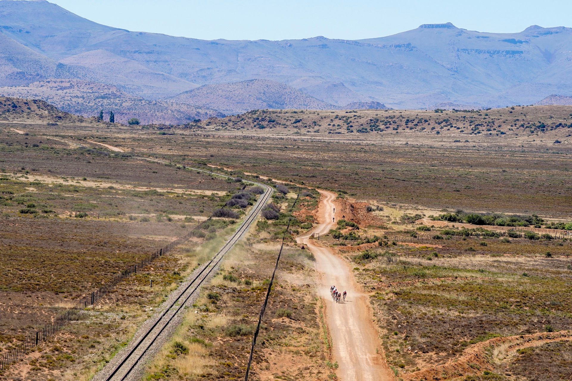 Riders on a gravel track beside a railway line across the Karoo plain, seen from above