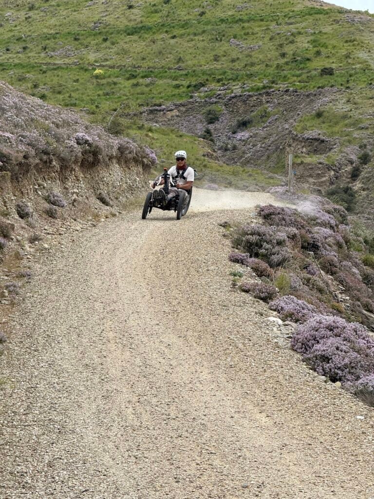 An adaptive rider on a Bowhead on a gravel road through purple heather in the mountains